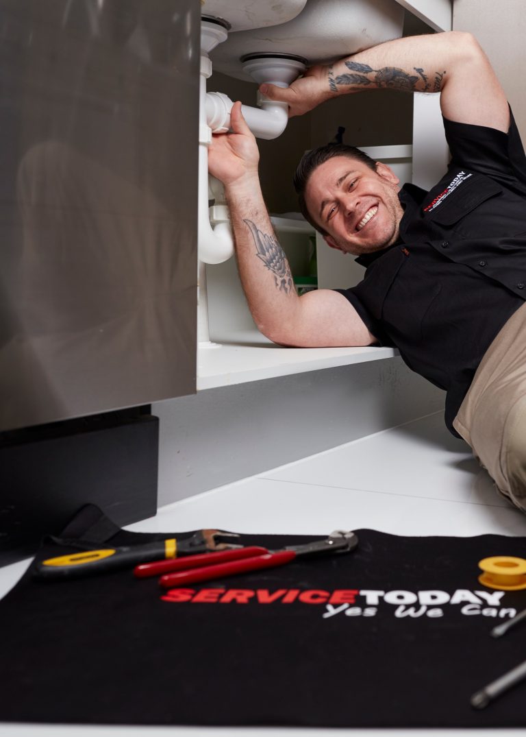 Plumber smiling while fixing a sink pipe with tools laid out on a mat.